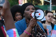 This still features a person with dark brown skin and a black head wrap who stands in the middle of a crowd on a city street and speaks into a megaphone. They are holding the megaphone in their right hand, and the megaphone has a heart-shaped trans flag sticker on it. The megaphone is mostly white, with the end having black edge pieces and the mouthpiece being red. The person is wearing an animal-print shirt with a light blue cardigan over it. On their ears, they wear silver feather earrings. They have two bracelets on their right wrist, one metallic pink beaded, and a silver one that does not fully connect around the wrist.
