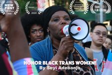 This poster features a still from the film with additional text imposed on top of it. The text at the top of the image is a series of badges from various festivals where the film was featured as an official selection. The text at the bottom of the poster is the title and creator, reading "Honor Black Trans Womxn! by Seyi Adebanjo." The still behind the text features a person with dark brown skin and a black head wrap who stands in the middle of a crowd on a city street and speaks into a megaphone. They are holding the megaphone in their right hand, and the megaphone has a heart-shaped trans flag sticker on it. The megaphone is mostly white, with the end having black edge pieces and the mouthpiece being red. The person is wearing an animal-print shirt with a light blue cardigan over it. On their ears, they wear silver feather earrings. They have two bracelets on their right wrist, one metallic pink beaded, and a silver one that does not fully connect around the wrist.