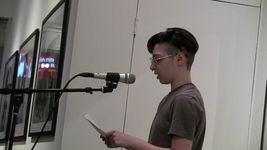 At a gallery, Sage Yockleson stands at a microphone reading from a piece of paper. They have light skin, short dark hair and glasses, and are wearing a plain t-shirt.