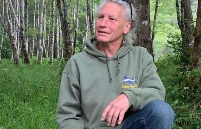 An image of Dina Boyer mid-inteview as she sits on the grass in a forest. She has light skin, gray hair pulled back, and wears a green hoodie with a graphic on it.