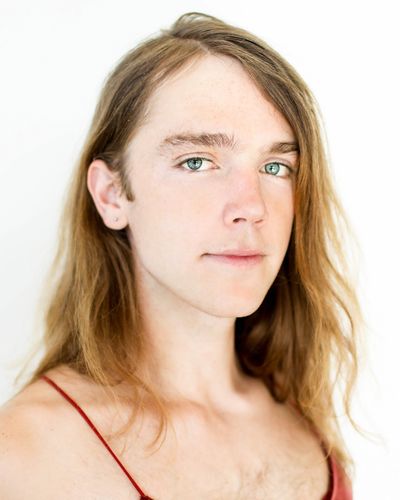 A soft and polished portrait of Zachary Clarence looking directly into the camera with a calm expression and standing in front of a glowing white backdrop. They have light skin and long, blond hair, and are wearing a thin-strapped tank top.