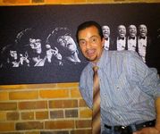 Boyd Kodak stands in front of a brick wall, smiling at the camera. He has medium tan skin and dark hair, and is wearing a striped shirt and a light brown tie. Behind him is a black-and-white photograph collage featuring various expressions of a singer mid-performance.