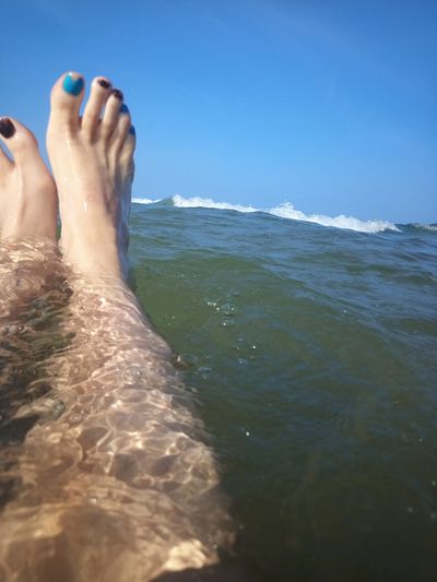 Two white feet floating in large body of water with a foaming wave. The sky behind is blue. The toenails are painted blue and brown.