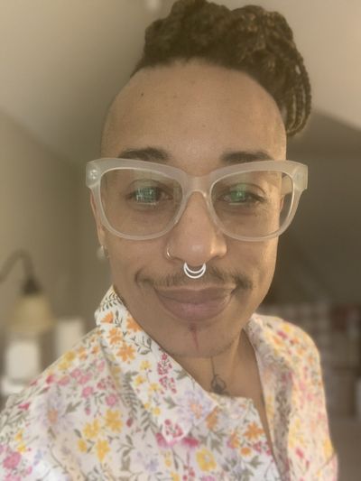 Close up face shot of a light-skinned person with a light mustache wearing clear glasses, a white septum ring, and braids on the top of their hair. The sides of their head are shaved close. They have a face tattoo of a red line down their chin, extending to a black line with a spider in their neck dip AKA jugular notch. They are wearing a buttoned-collar white shirt with pink, yellow and orange small flowers. The background is blurry.