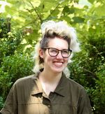 Candid portrait of Rainn Mowris in a leafy-green outdoor setting. Rainn is a white nonbinary person with a mop of brown-and-blonde hair perched atop their head and a large pair of round glasses on their face. They wear an olive jacket and look just past the camera with a smile.