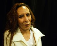Alexus Young is a First Nations person with medium brown skin and long, wavy brown hair. She is standing in front of a black background, smiling slightly. She is wearing a white jacket over a white top and large dangling earrings.