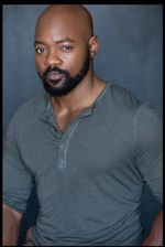 Professional studio portrait of Brian Michael Smith from the torso and above. Brian has dark skin, dark facial hair and a bald hairstyle. He is wearing a earthy blue coloured long sleeve shirt with his forearms exposed.