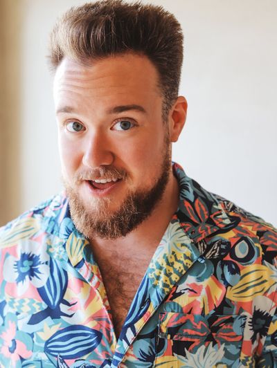 Bright and summery portrait of Zeke Smith with slightly widened, animated eyes and open smile. Zeke has light, tanned skin, medium short light brown hair that is styled upward, and light brown facial hair. He is wearing a colourful hawaiian shirt with chest hair exposed.