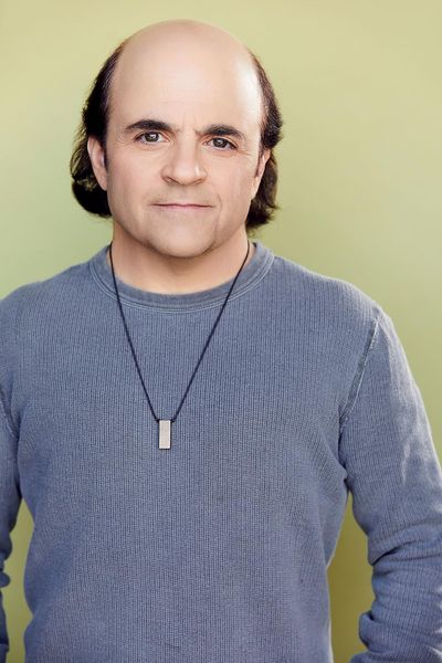 High quality professional studio portrait of Michael D. Cohen standing with his arms in his pockets against a solid light green backdrop. Michael has light skin, dark brown hair and eyes. He is wearing a dark blue long sleeve shirt with a minimal dark necklace.