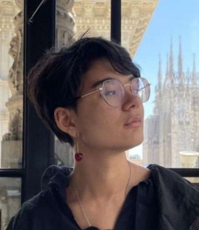 Mira De Koven seen in profile before a window to European-looking buildings, a poetic expression on their face. Mira is a nonbinary and mixed race person with short dark hair, big round glasses, and a glass cherry earring.