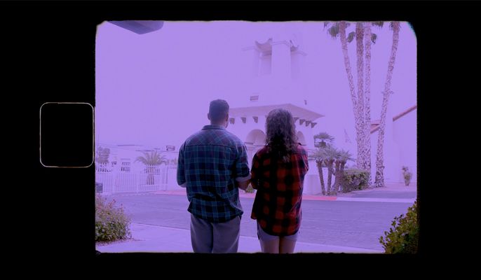 Kai & Love together in Love's imagination. Both wear brightly-colored plaid shirts and face away from camera, towards a white building surrounded by palm trees. There is a purple haze over the image, and the colors of the surroundings are washed out by bright daylight.