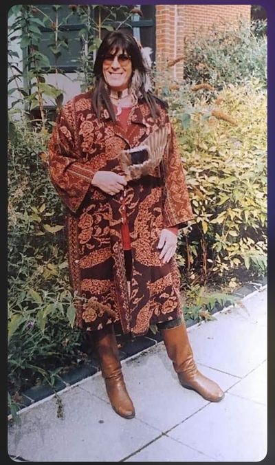 NapéWastéWiñ grins at the camera. She has black hair past her shoulders with bangs. She wears sunglasses, a long umber back with a yellow pattern on it, and brown boots. She holds a large brown bird wing in one hand.