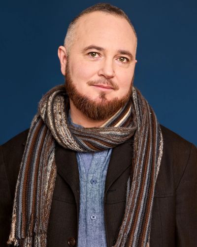 Studio portrait of Hanlon Uafás-Álainn, a trans man with light skin, a bilbo beard, and short auburn and white hair. He wears a knit scarf, a denim button-up and a blazer.