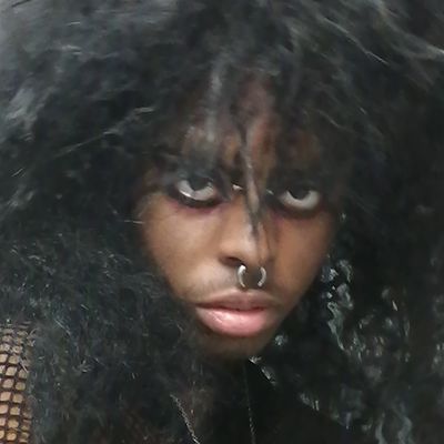 Close cropped photo of Thorne Brown, a Black trans man. He has heavy eye-make-up, a septum piercing, and a slight mustache. His hair is styled in a blow-out annd he wears mesh and a reflective fabric like pleather.