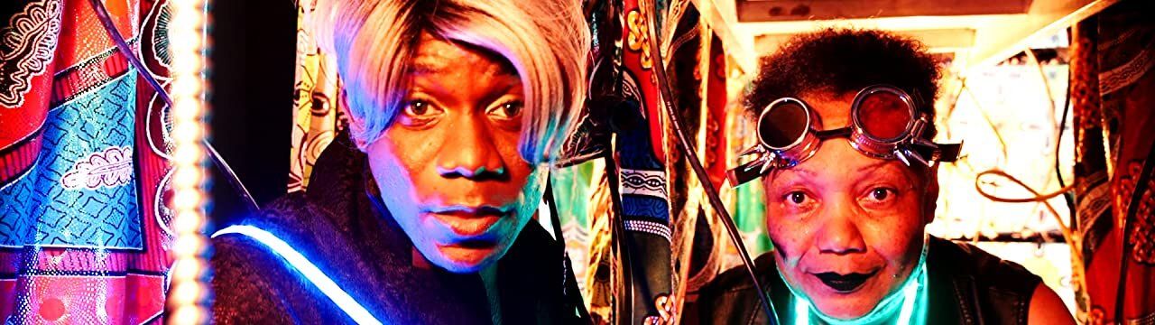 Two characters from "Ancestors, Can You Read Us? (Dispatches from the Future)" stare directly at the camera. The person on the left (played by Ravyn Wings) has a blonde bob, medium brown skin, and wears a black jacket with sash made of LED lights, while the person on the right (played by Gloria Swain) is slightly shorter with a short blow-out, light brown skin and round cyberpunk glasses. They appear to be in a tent made of kitenge or other African-style batik with some hanging wires.