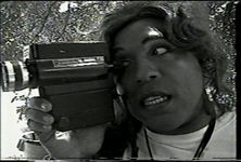 Black-and-white film still of Vaginal Davis peering through a video camera. Vaginal Davis is a Black and Hispanic person with wavy long hair wearing a t-shirt.