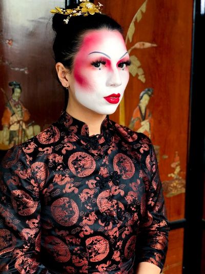 Kendall looks at the camera with slightly parted lips. She is wearing heavy makeup with a white base, red lip, thin arched brows, long eyelashes, and bright red blush and eyeshadow. She has dark hair styled in an bun and wears a garment that is tailored to fit of a qipoa but has cloth buttons symetrically down the front in a style common in tang shirts.