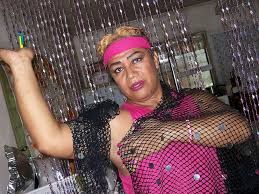 A person wearing matching hot pink headband, top, and lipstick with a mesh shawl drapped over them. They have short blonde hair, light skin, and a pose suggestive of dancing. They are in front of a  glittery beaded curtain.