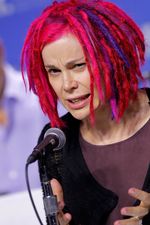 Lana Wachowski seen at a dutch angle from the shoulders up as she speaks emphatically into a microphone. Lana is a white trans woman with shoulder-length dreadlocks coloured a dramatic hot pink, with highlights in several shades of purple and pink. She wears a plum shirt beneath a black blazer.