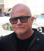 Grainy close-up selfie of Mickey R. Mahoney in a parking lot on a sunny day. Mickey has light skin, is bald with a thin greying mustache and chin beard, and wears dark sunglasses and a denim jacket over a black shirt.