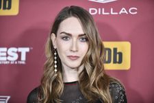 Red carpet headshot of Jamie Clayton with raised eyebrows and a faint smile. Jamie is a white trans woman with dirty blonde hair and blue eyes. She wears a dangling string of pearls as an earring and a dark nylon dress accentuated with clusters of little metal flower petals.