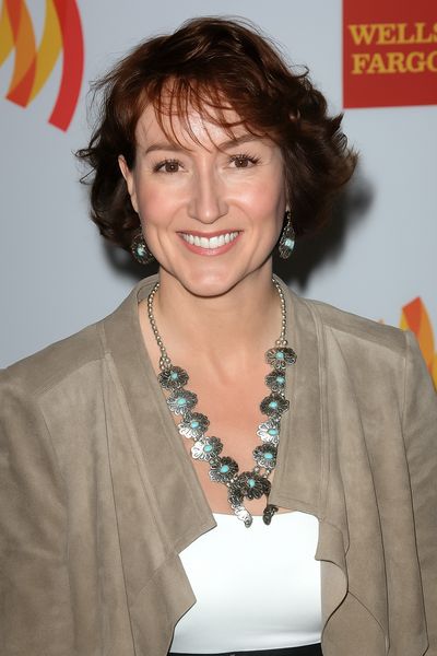 Red carpet headshot of Lydia Nibley, a light-skinned person with wavy chin-length auburn hair and brown eyes. She wears a white top, leather jacket, and silver & topaz jewelry.