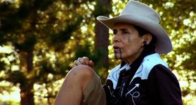 Still of a person resting outside. They have light skin, dark hair, and facial tatoos. They wear a wide-brimmed rodeo-style hat and western button-up shirt.