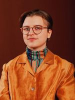 Cassidy Civiero is shown from the chest up in front of a brown studio background. He has round glasses and short brown hair stylishly parted to the sides. Cassidy is wearing a shiny orange suit over a brightly striped multi-color turtleneck.