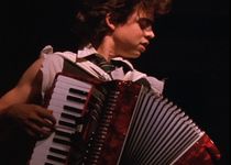 Geo Wyeth plays an accordion in a black void, his head seen in profile as he closes his eyes and loses himself in the performance. He has a mop of curly brown hair, light skin, and wears a white shirt with torn sleeves revealing his arm muscles.