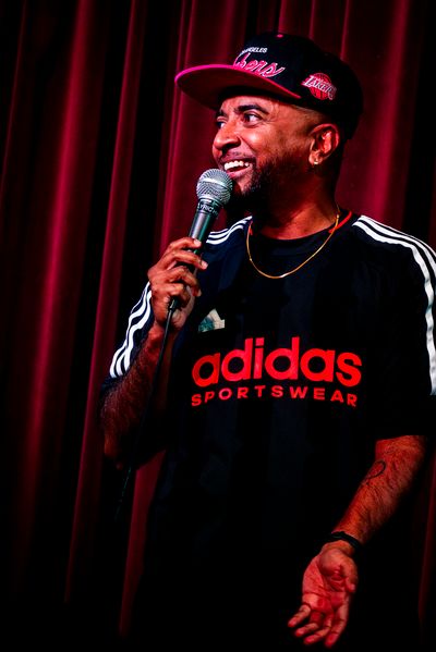 A brown trans guy is holding a mic, doing stand up and has a cheeky smile. He is wearing a black adidas shirt, a thin gold chain, and a pink lakers' baseball cap.