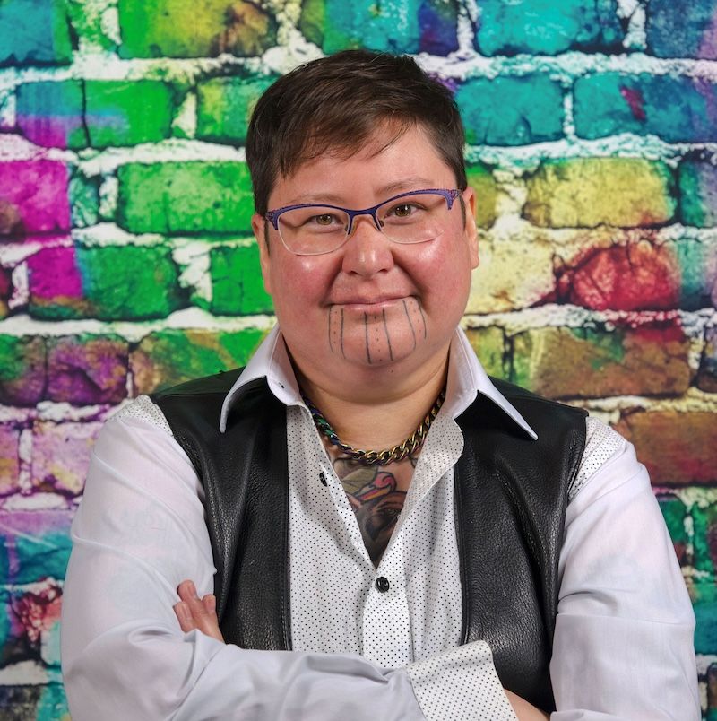 Theo Jean Cuthand wears glasses, has short hair, and is a light-skinned                         person of Plains Cree and Scots descent. He has facial tatoos on his chin of dots and vertical                         lines. He is wearing a white button-up with a black leather vest and is standing in front of a rainbow-coloured brick wall