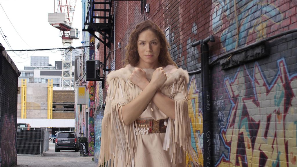 Film still from Woman Dress. An Indigenous person with long wavy brown hair past, wearing a light-brown fringed leather dress lined with fur and a beaded belt stands looking at the camera with their arms forming an X in front of them, their hands closed into fists. They are standing in an urban alleyway beside a colorful graffitied brick wall.