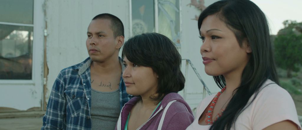 Still from Drunktown’s Finest (2015). Three characters look at something off-screen.The person on the far left has light tan skin with a blue and white flannel and a grey muscle t-shirt underneath. They have a buzz cut with black hair and tattooed words over their chest that cannot be made out in the image. The person to their right has shoulder length black hair, tank skin, a dark purple zip up. The person on their right has long straight black hair with tan skin a white short sleeve top and a red and blue necklace around it.