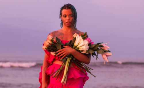Tourmaline, a brown-skinned person with dark, wet, shoulder-length hair, stands in front of a sunset in a pink ruffled dress, her left foot on the beach and right foot slightly lifted. She holds a bouquet of tulips in her left hand, holding up her dress with her right hand. The background is a blurred out beach with the waves crashing in. They have a tattoo on each shoulder unable to be made out.