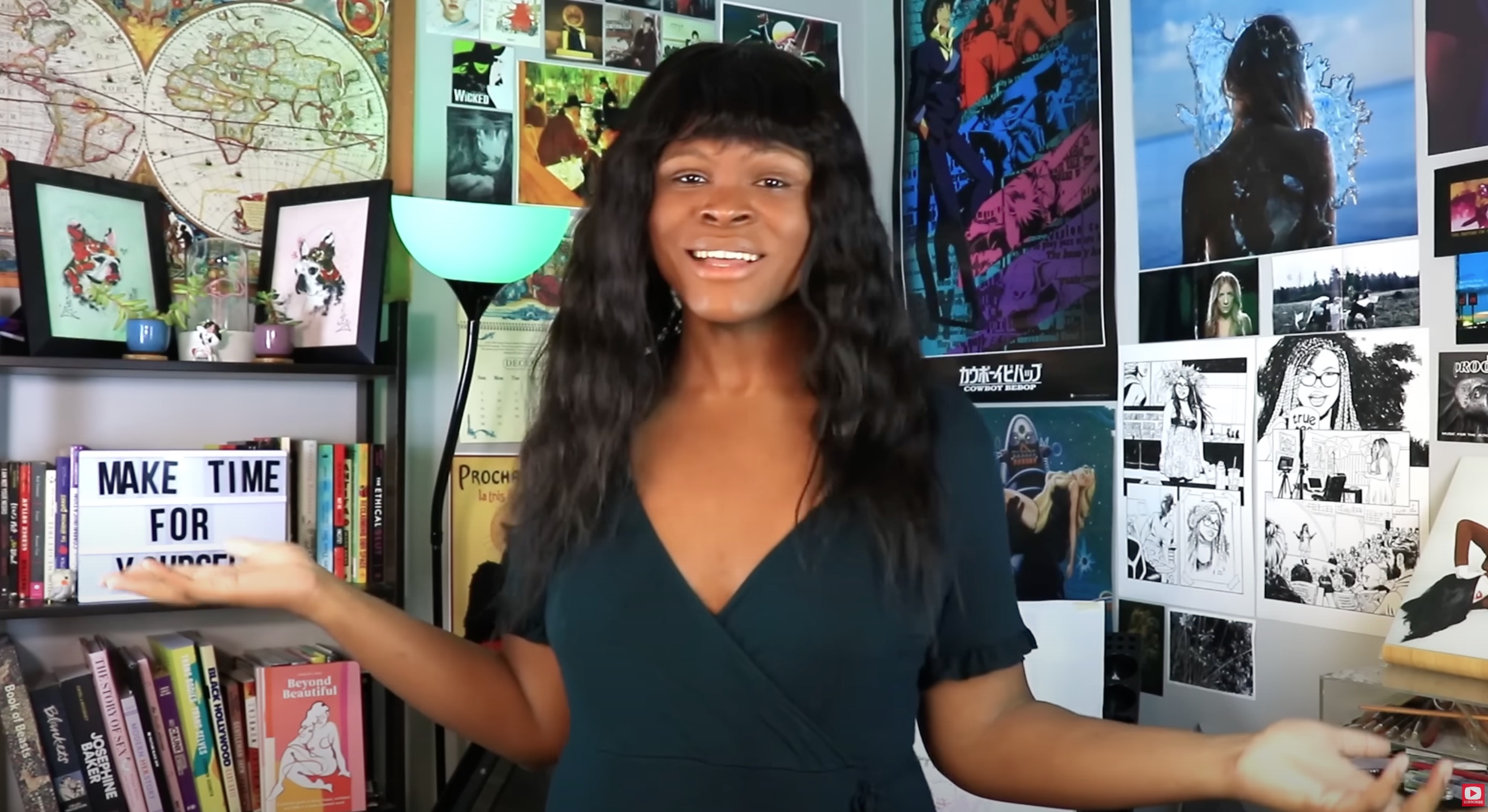 Kat Blaque, a Black person with medium skin and dark wavy hair with blunt bangs, stands in front of a heavily-postered wall with her arms raised in a questioning shrug and wearing a navy blue v-neck short sleeve top.
