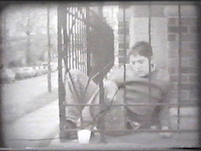 Black and white still from The Man from Venus (1999). Diamond sits between an iron fence and a brick wall, his expression cast downwards.